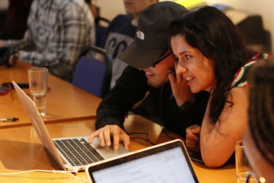 Two young people sitting infront of a laptop, both listening to the same set of headphones